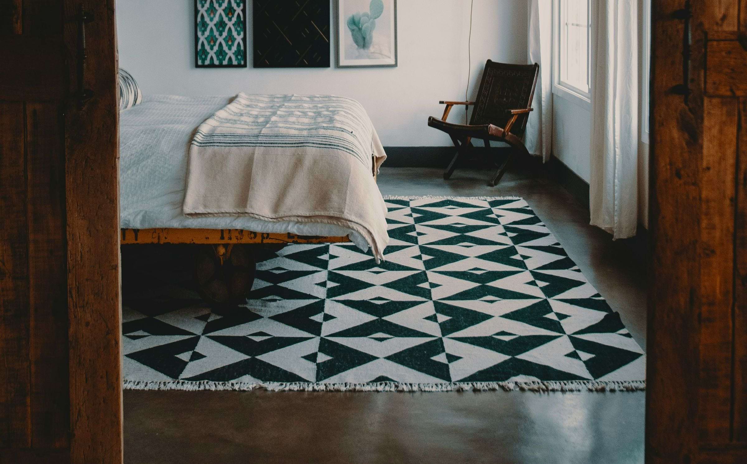 A bedroom with a dark floor and a black and white geometric rug, which extends out from the end of the wood-framed bed.