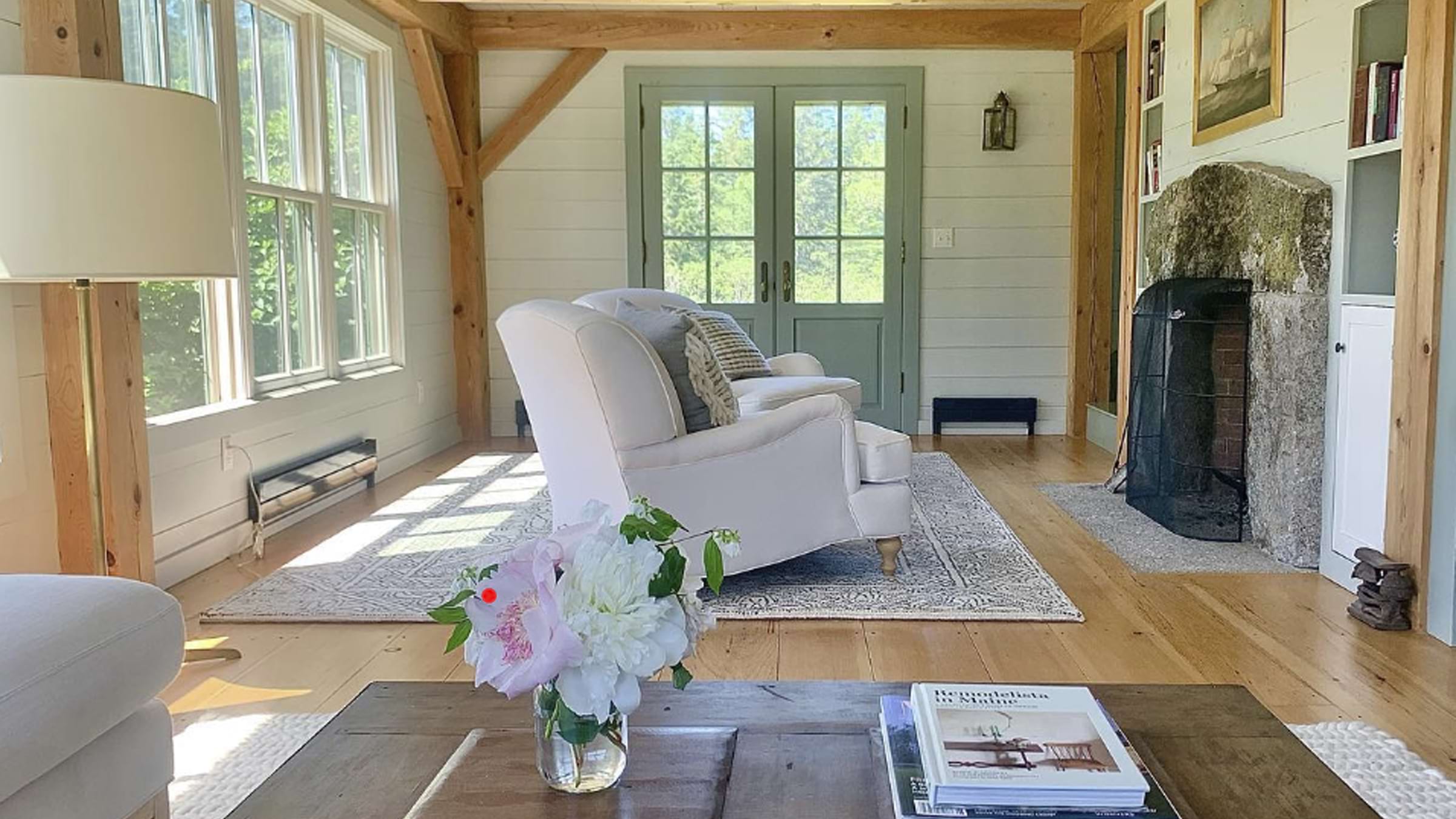 The interior of home with white chairs in the center of the room on a rug. A table with books and a vase of flowers is in the foreground and the room features a lot of natural light.