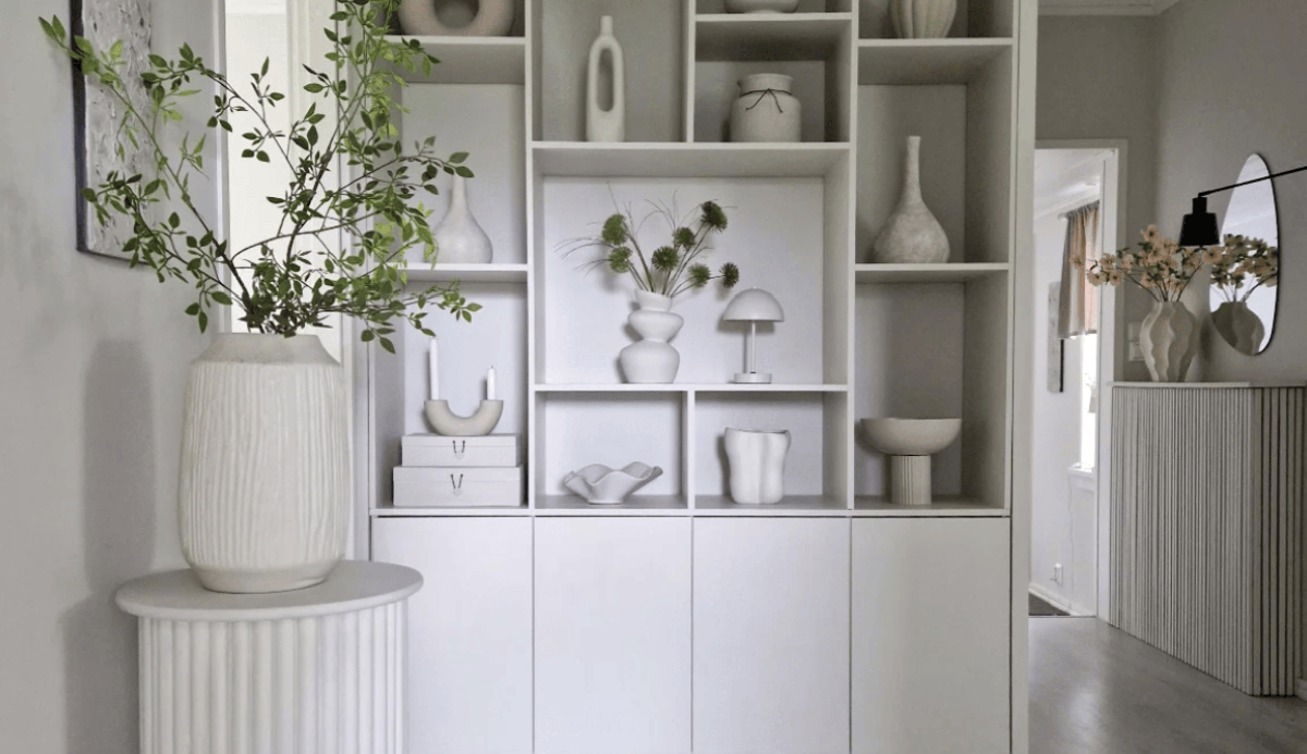 Corner of a room in a house that shows a beige shelf built into the wall and a plant in an off white pot sitting on an off white table. In the background is a hallway with a mirror, lamp, and table.