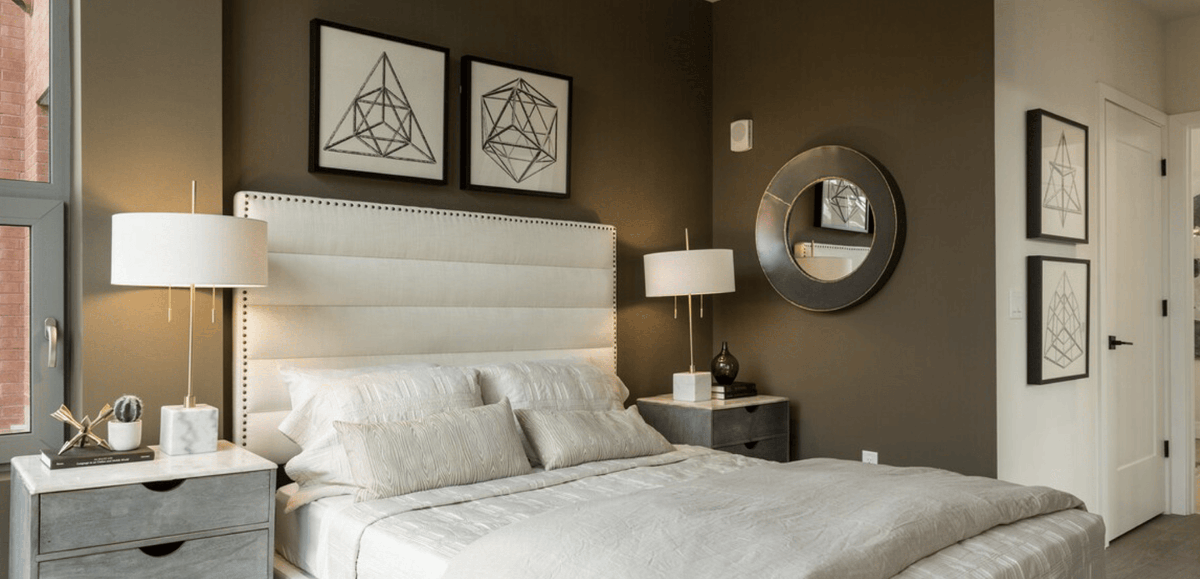 A bedroom with dark brown walls and matching mirror with a white bed, lamps, and framed art,