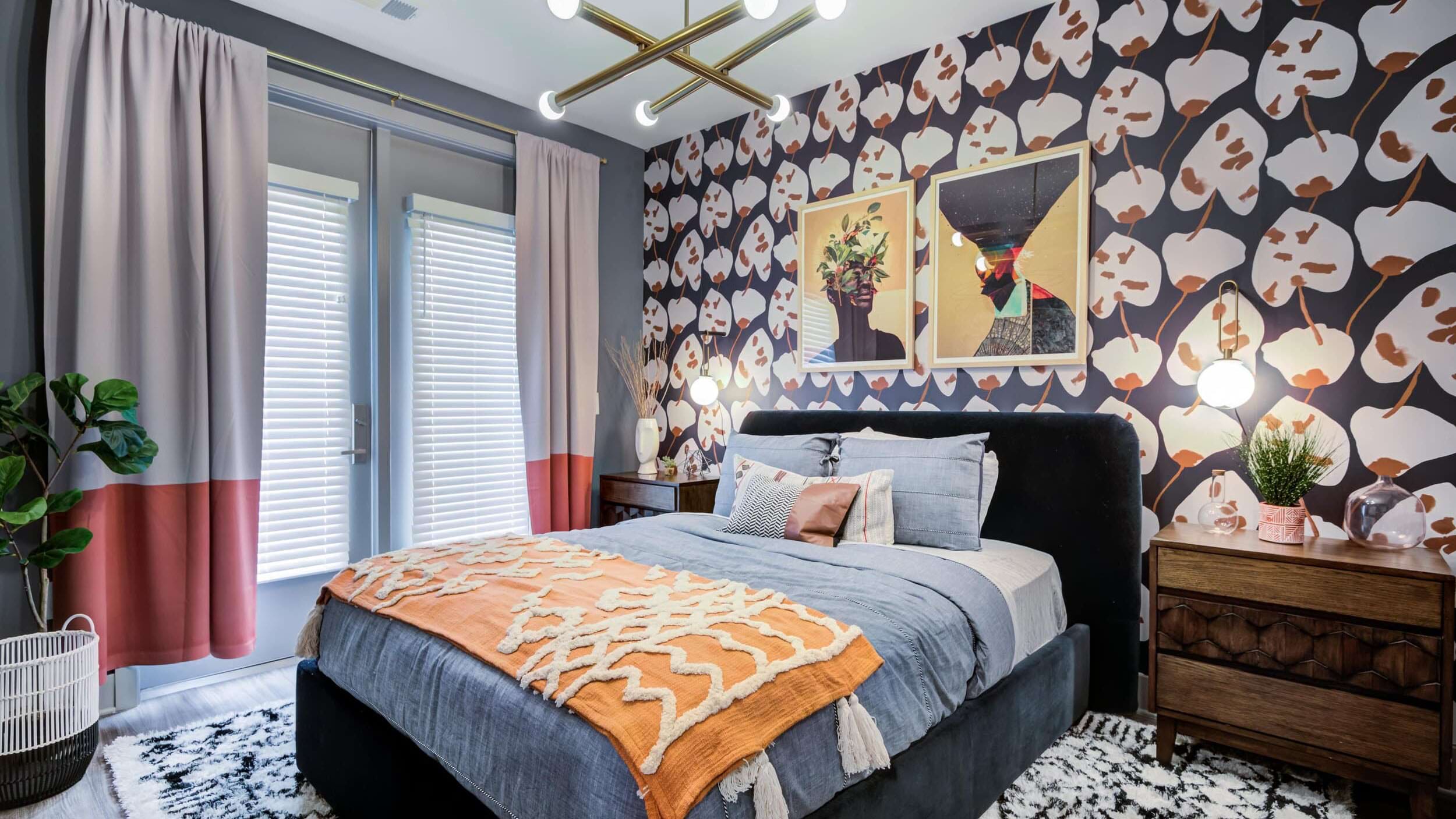 Interior of a bedroom with a patterned wall, large windows with color block curtains, and a bed with a blue blanket layered with an orange throw. There is a gold light fixture on the ceiling and large square framed art on the patterned wall.