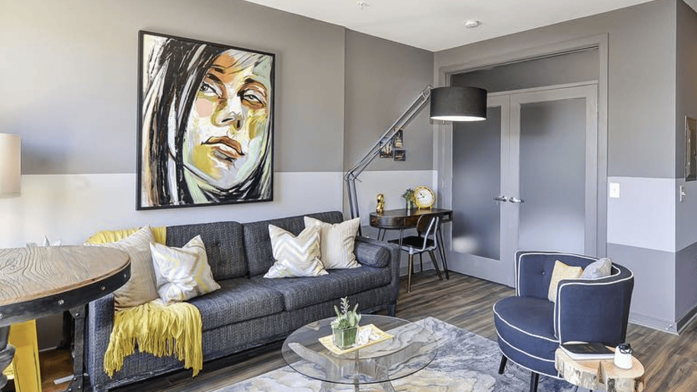 A living room with gray and white walls featuring a large square piece of art - a line drawing of a female with dark hair. On the same wall as the painting is a gray couch next to a large interesting lamp and a small desk and chair. A glass coffee table and blue chair are pictured in the foreground of the image.