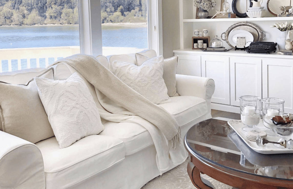 Living room with a view of lake out the back windows. In front of the windows are a white couch and blankets, a white shelf built into the wall, and a brown wood coffee table.