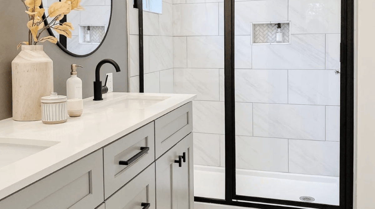 A bathroom with off white marble counter tops, vase, and soap dispenser, gray cabinets and wall, and black fixtures and shower door frame.