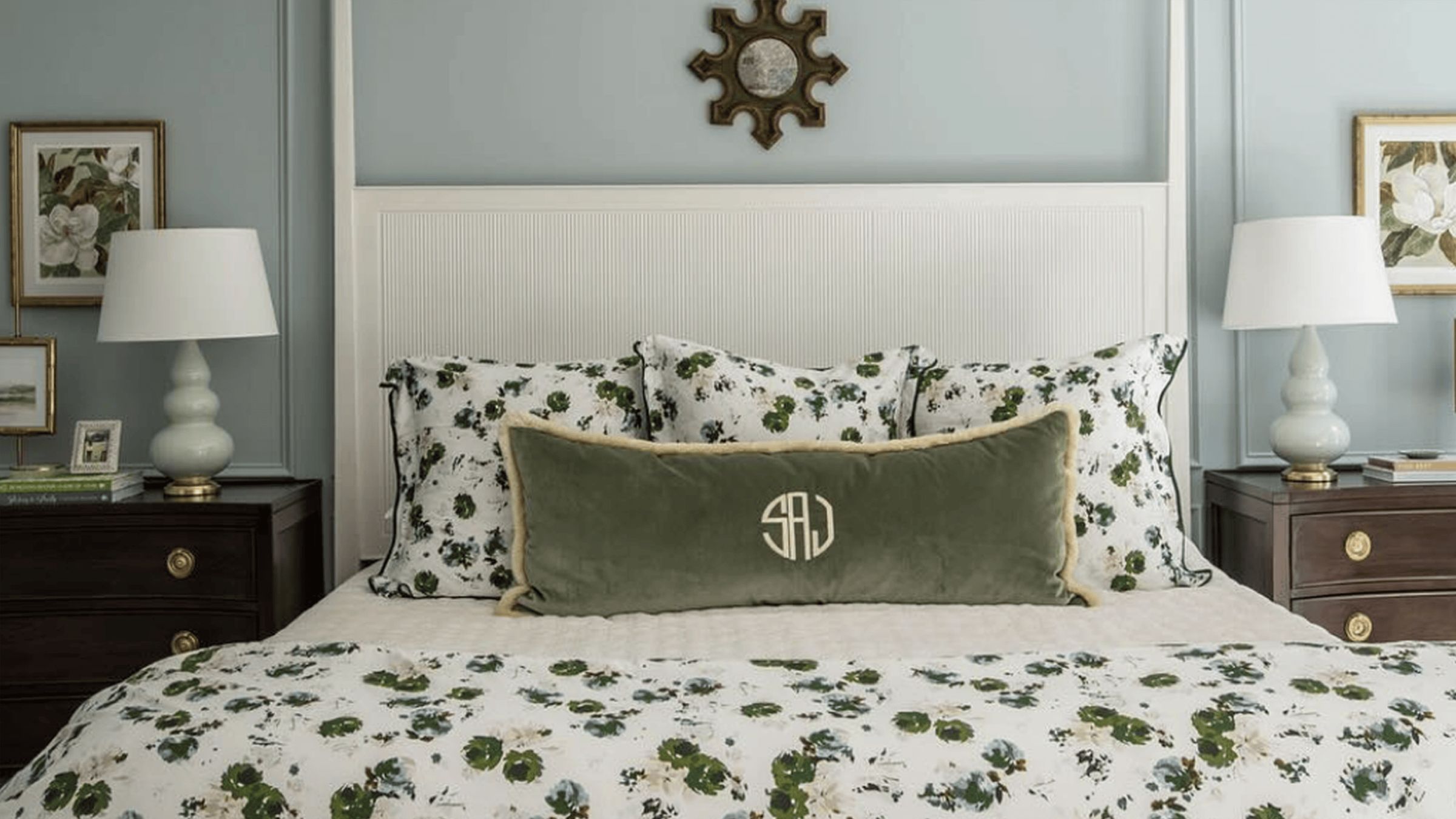 A large bed with a white headboard and green and white bedding is centered on a contrasting green wall between two side tables, lamps, and framed art. In the center of the bed is a dark green, velvet pillow that is monogramed.