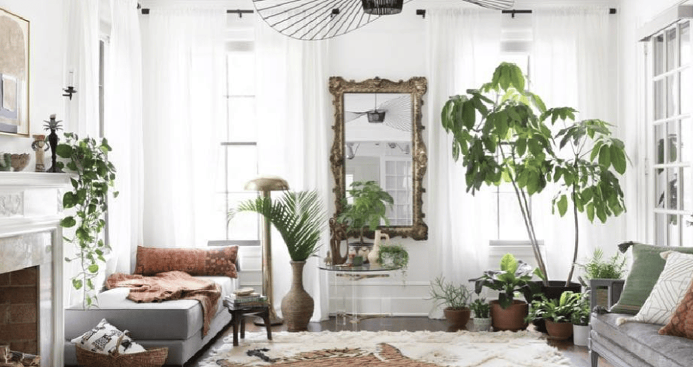 Interior of a large living room with white walls and gray furniture. Centered in the image is the back wall of the rooms with multiple green plants at varying heights and a large rectangle mirror with a brass frame.