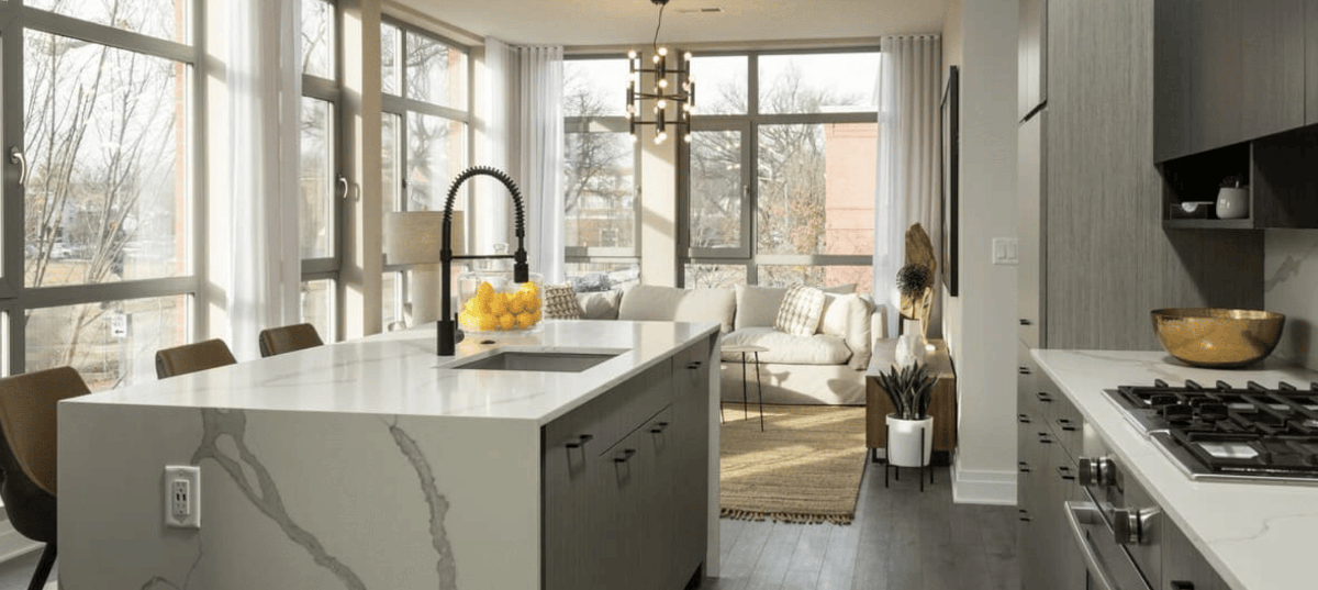 Neutral colored kitchen with gray walls, floor to ceiling windows, and light colored marble countertops. A bowl of lemons sits on the light colored marble countertop of the kitchen island. An off white couch and lamp are seen in the background.