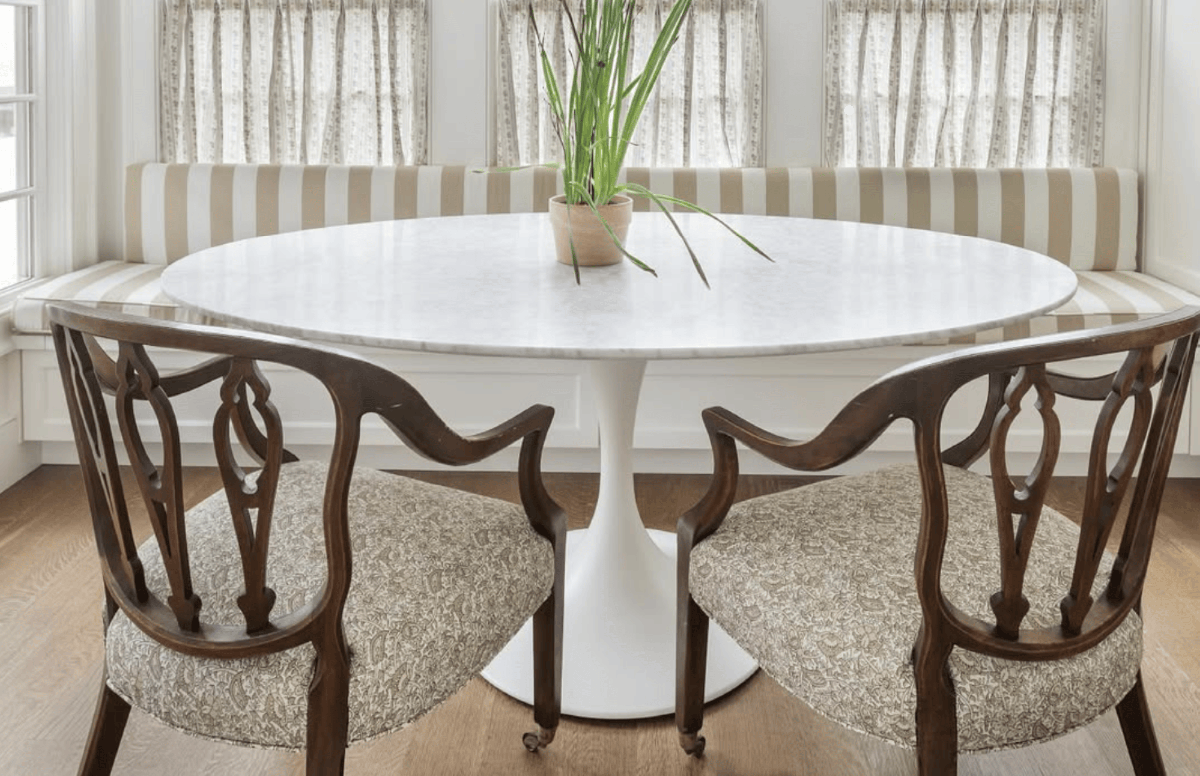 A dining area in a home with a white table holding a plant, brown and white striped fabric bench, and two brown chairs.