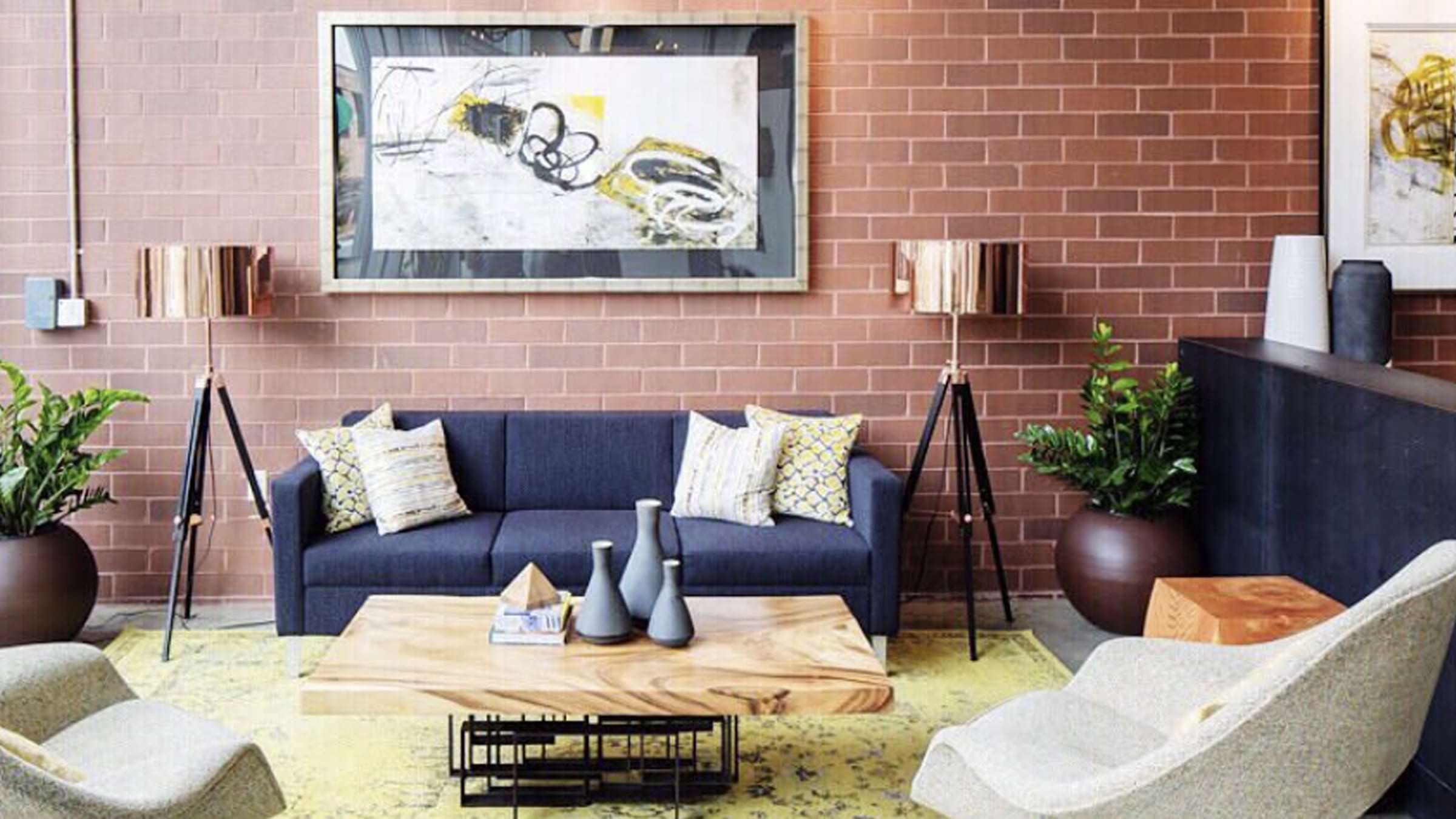 Living room with a red brick wall featuring a large peice of framed art. A navy blue couch with light yellow pillows is centered below the art and a coffee table is n the foreground. A floor lamp and large plant are to the right of the couch