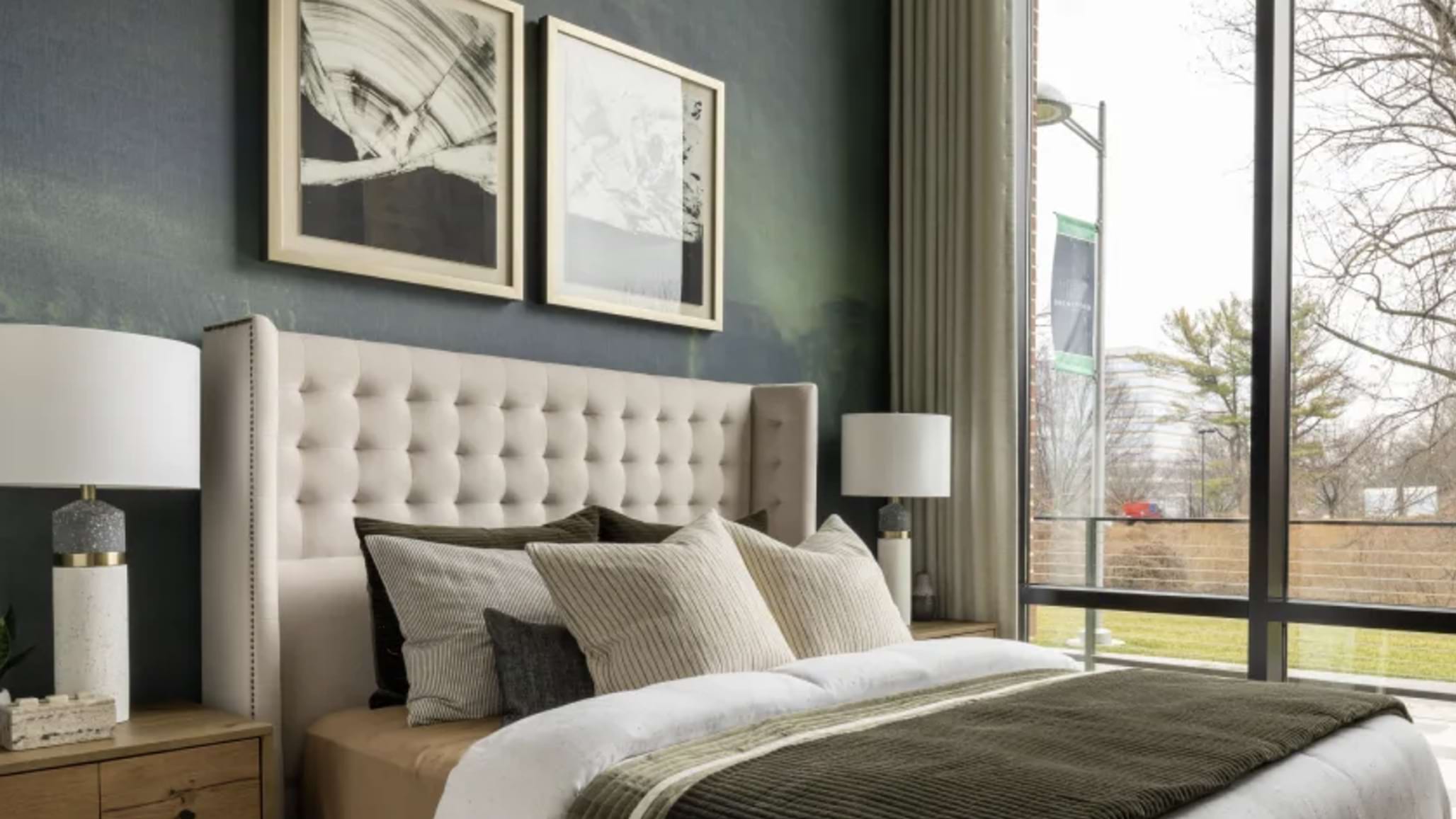 A bedroom with tall ceilings and a dark green wall on one side. On the wall is a bed with a fabric headboard and multiple decorative pillows next to a table with a lamp. 