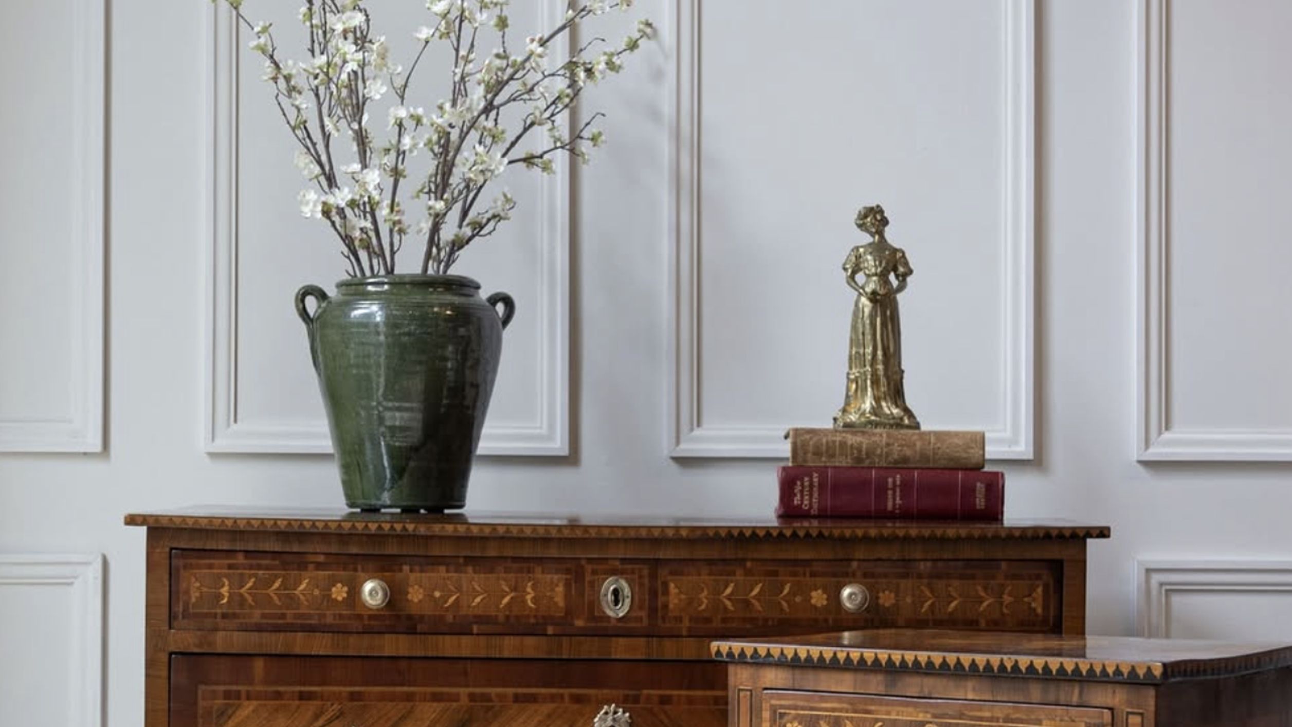 A wooden bureau with a large ceramic vase holding dried flowers and a brass statue sits on a stack of books.