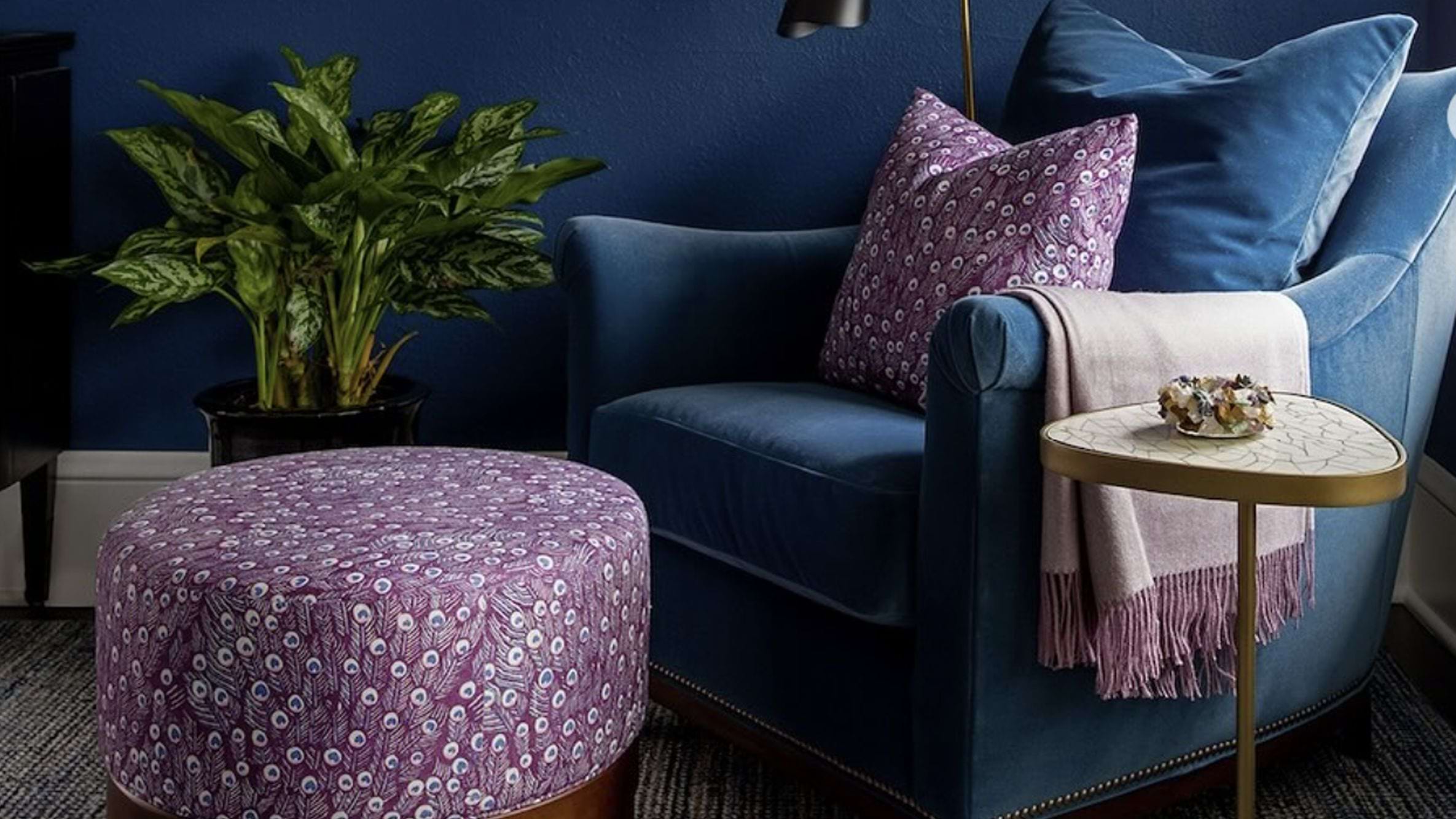 A dark blue velvet couch against a coordinating blue accent wall with lavender details including a pillow and ottoman.