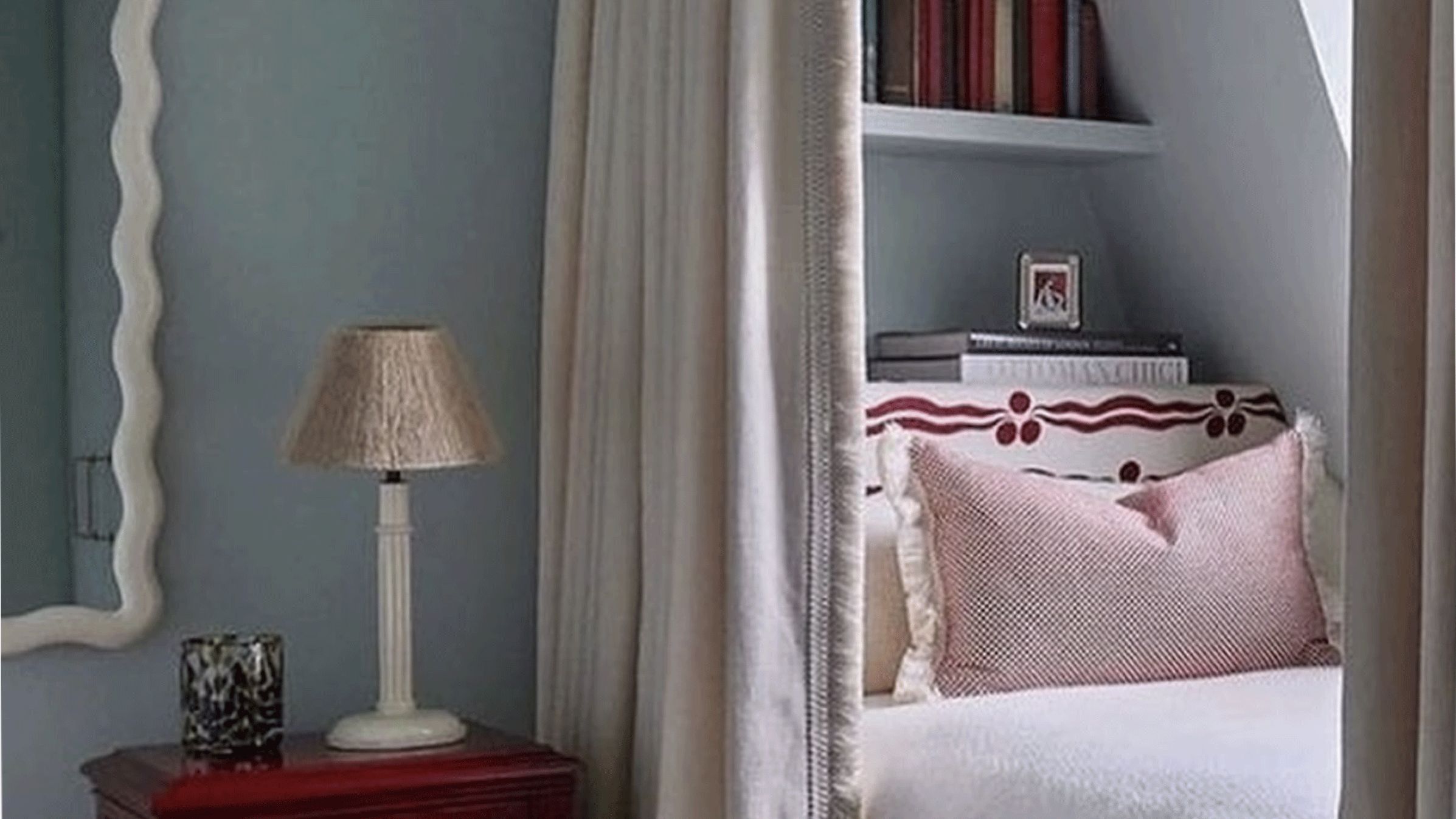 A bed is built into a space and there is a vertical shelf behind the head of the bed. A curtain hangs to the left of the bed and there is a small dresser with a lamp.