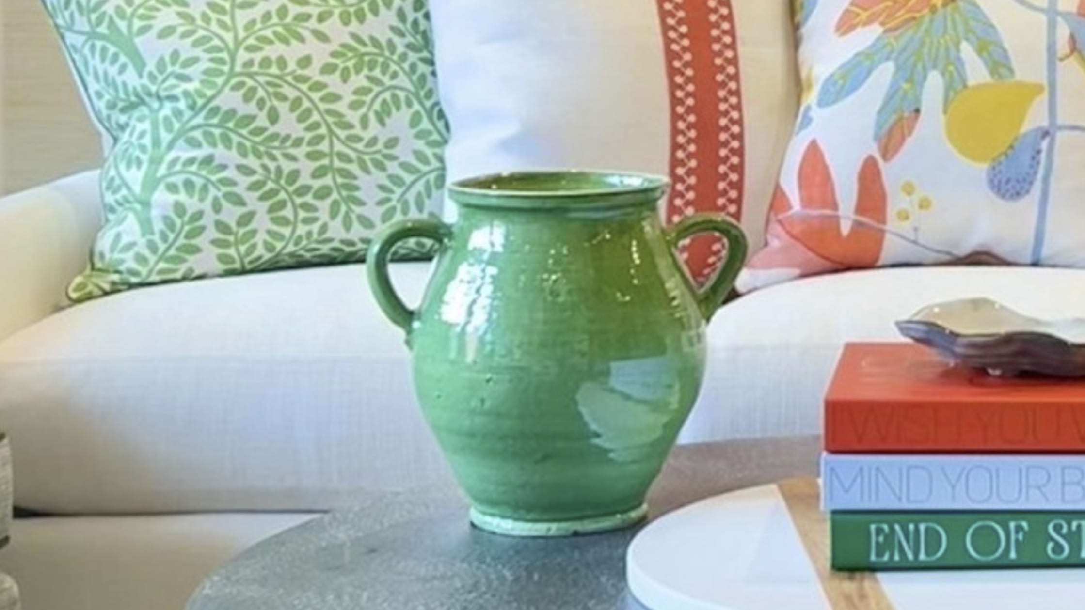 A green ceramic vase sits on a table in front of a bright couch with pillows that are the same shade as the vase.