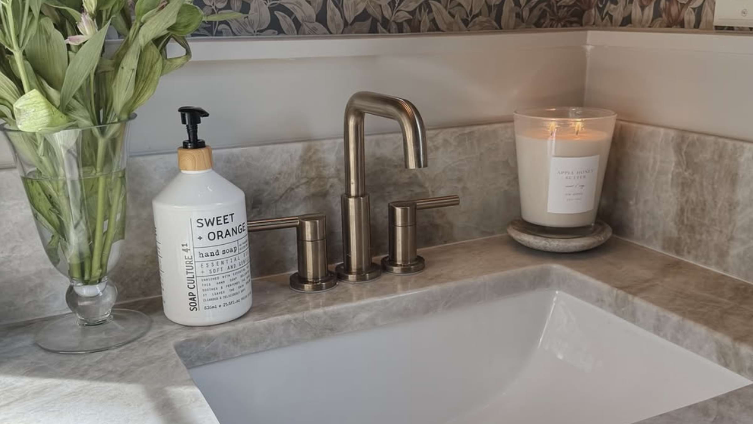 A bathroom sink with a vase of flowers, a soap dispenser that reads "Sweet Orange," a faucet, and a lit candle.